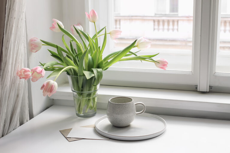 A white table and window with a cup of coffee and pink tulips in a vase