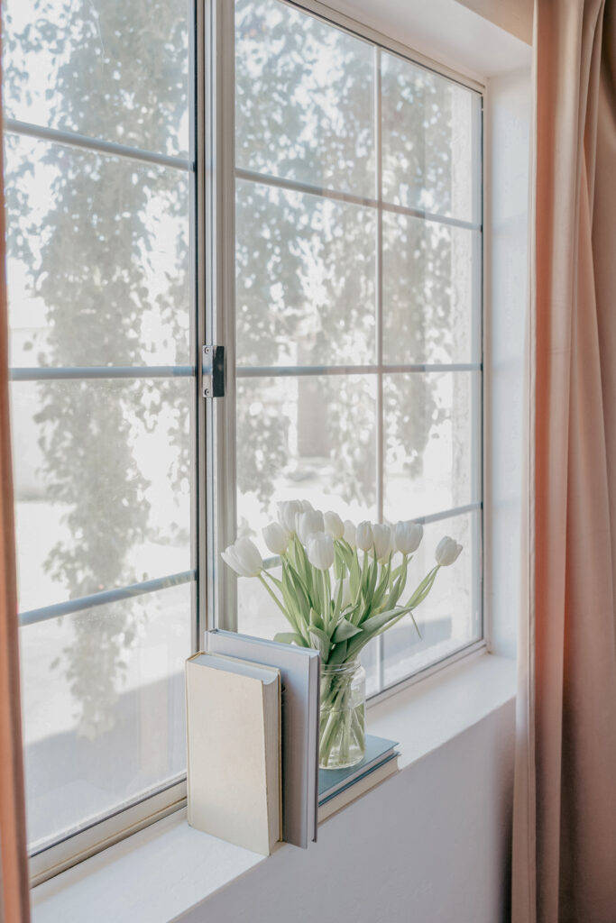 A white window frame with peach-coloured curtains and white tulips in a vase